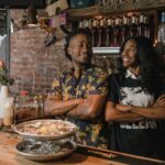 Two self-employed small business owners standing behind their bar, smiling confidently with arms crossed.