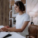 Young self-employed woman working on laptop at home office desk, researching health insurance options as a 1099 contractor