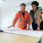 Construction worker in orange safety vest reviewing blueprints with a colleague, representing the need for proper health and accident insurance coverage for contractors and trades workers.