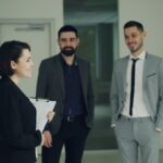 Small-business team of four colleagues in business attire talking and smiling in an office hallway while one holds a clipboard