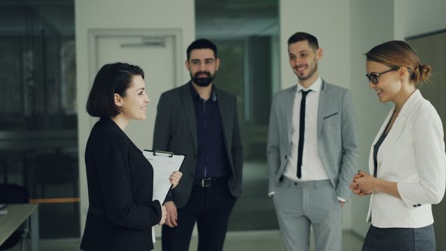 Small-business team of four colleagues in business attire talking and smiling in an office hallway while one holds a clipboard