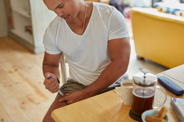Man using an injectable medication pen on his thigh while sitting at a kitchen table with coffee