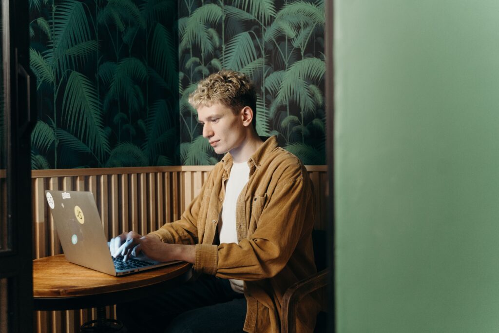 Young man in a tan corduroy shirt typing on a laptop at a round wooden table in a booth with palm-leaf wallpaper.