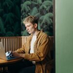 Young man in a tan corduroy shirt typing on a laptop at a round wooden table in a booth with palm-leaf wallpaper.
